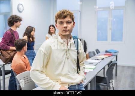Young man with a backpack standing in a university classroom, with diverse students gathered, preparing for a lesson, representing education and campu Stock Photo