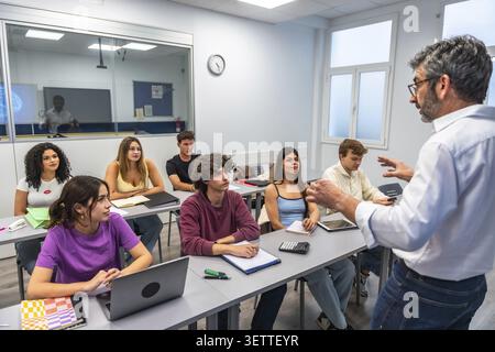 Professor giving an engaging university lecture to attentive students in a modern classroom, taking notes and using laptops and tablets during interac Stock Photo