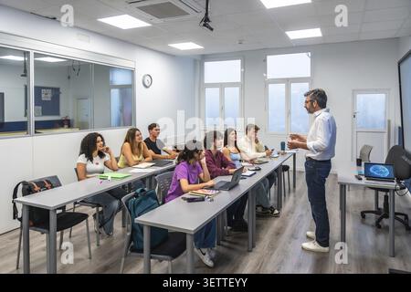 Teacher at front of bright classroom explaining lesson to diverse young adult students seated with laptops, engaging in discussion and attentive moder Stock Photo