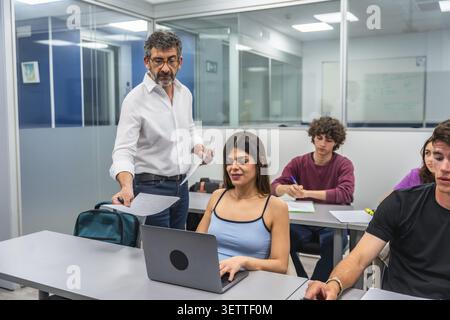 Professor interacting with diverse students in a bright school classroom, helping a young woman using a laptop for her studies while others are taking Stock Photo
