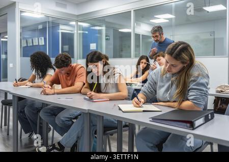 Adult students sit at desks in a bright modern classroom, focused on writing notes and taking an exam while a male teacher supervises their concentrat Stock Photo