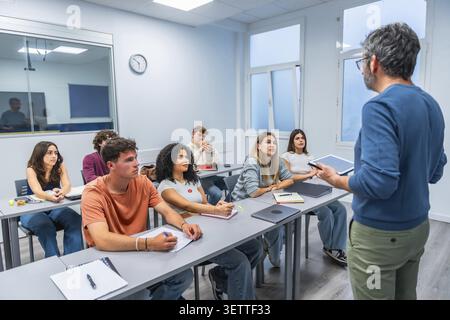 Teacher leading an engaging university lecture as diverse students listen, take notes and use digital devices in a modern classroom, promoting collabo Stock Photo