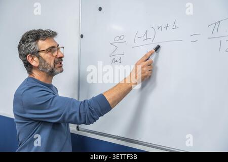 Male professor teaching advanced mathematics, pointing to complex equations on a whiteboard during a focused classroom lecture or online course, expla Stock Photo