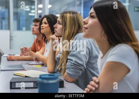 Young diverse students sitting at desks in a modern classroom, actively listening and engaging with educational content during a university lecture or Stock Photo