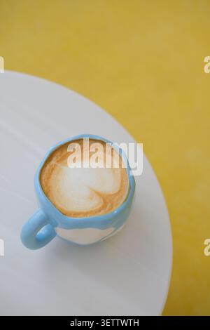 Blue ceramic cup with creamy latte foam on white table against yellow background Stock Photo