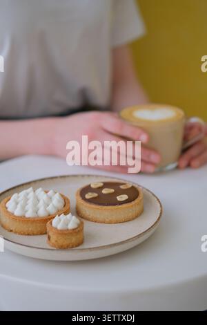 Hand holding latte beside pastry plate with assorted tart desserts on table Stock Photo
