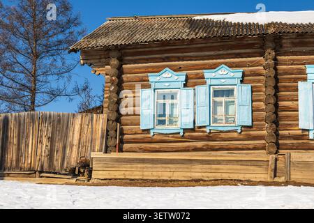 View of a traditional wooden log house with bright blue window frames and shutters, set against a clear blue sky with snow on the ground in Baikalskoy Stock Photo