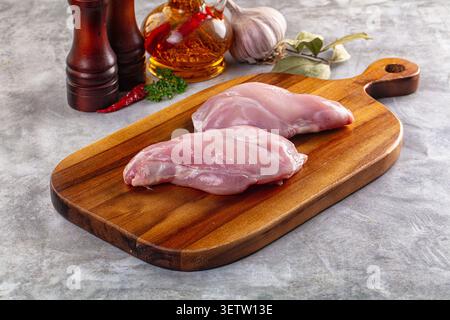Raw rabbit thighs on rustic wooden cutting board. Background includes pepper mill, garlic, chili, bay leaves, oil bottle. Soft natural lighting, warm Stock Photo