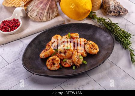 Grilled shrimp served on matte black plate with fresh rosemary sprigs lemon pink peppercorns. Coastal theme with seashells on marble background. Warm Stock Photo