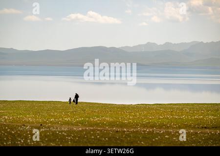 A distant silhouette of a man holding a small child's hand as they walk along the grassy shore of Son Kul Lake in the Kyrgyzstan mountains, with a cal Stock Photo