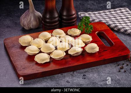 Raw pelmeni dumplings arranged on red wooden cutting board. Garlic bulb, pepper mills, parsley, checkered cloth on dark stone background Stock Photo