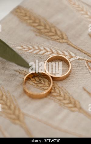 A close-up, elegant shot of two simple gold wedding bands resting on a light beige textured linen cloth decorated with delicate golden wheat embroider Stock Photo