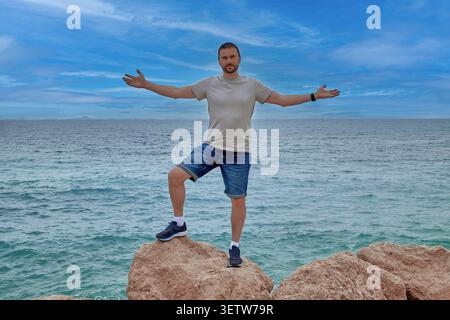A young man stands on a stone against the background of the sea with his arms outstretched Stock Photo