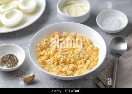 Crumbled hard boiled egg yolks in a bowl, garlic, mayonnaise and seasonings, prepared as filling for stuffed eggs on a gray textured surface Stock Photo