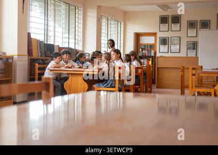 Group of primary school students gathered around a table in a school library, reading and learning together under teacher supervision. Stock Photo