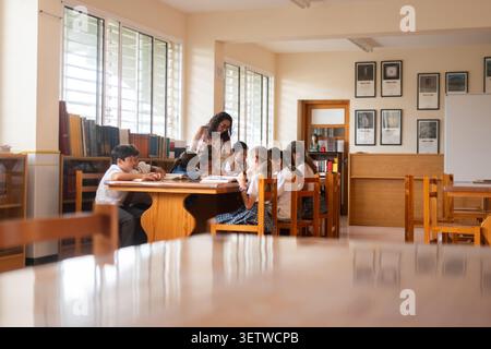 Group of primary school students gathered around a table in a school library, reading and learning together under teacher supervision. Stock Photo