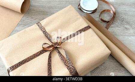 Gift box wrapped in brown kraft paper with leopard print ribbon and a roll of matching wrapping paper on a wooden surface Stock Photo