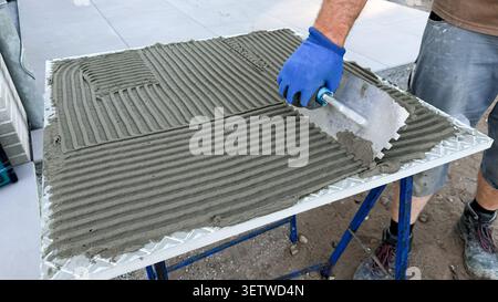 Male construction worker applying cement mortar with trowel on a flat surface at a construction site, blue gloves and tools visible in the background Stock Photo