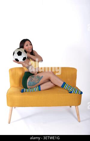 A young woman sitting on a yellow sofa while holding a soccer ball in a minimalist studio setting. She wears sporty clothing and colorful striped sock Stock Photo