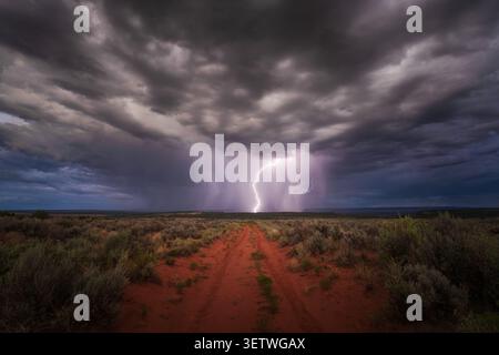 View of a powerful lightning strike illuminating dark storm clouds over a red dirt road and desert landscape in Kanab, Utah, United States. Stock Photo