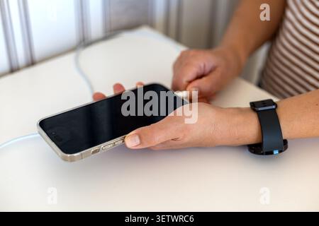 Hands plugging charging cable into smartphone. Close-up of hands connecting a white charging cable to a smartphone on a white table, with a black Stock Photo