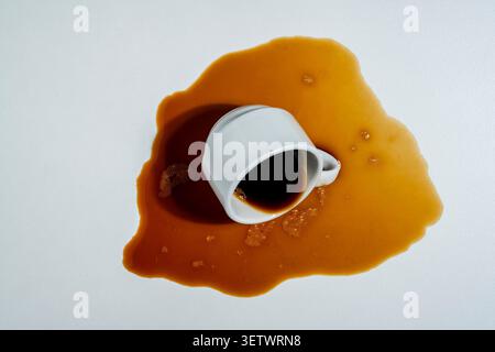 Spilled coffee from overturned white cup on light background, close up. Top view of a spilled puddle of dark espresso coffee with a fallen white ceram Stock Photo