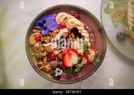 Vibrant acai smoothie bowl topped with sliced banana, fresh strawberries, granola, almonds, and edible flowers. Stock Photo
