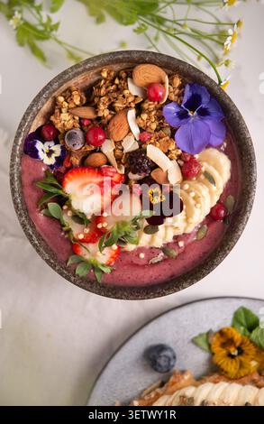 Vibrant acai smoothie bowl topped with sliced banana, fresh strawberries, granola, almonds, and edible flowers. Stock Photo
