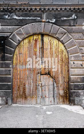 Old wooden door with rust and wear under stone arch. Stone wall with weathered texture. Ground covered in gravel Stock Photo
