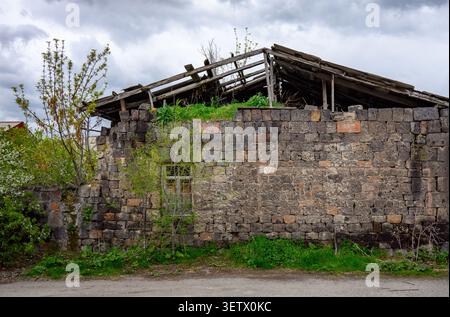 Old stone ruin with broken roof. Vegetation grows through cracks. Cloudy sky above. Small tree beside wall. Grass covers base Stock Photo