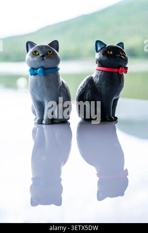 Chiang Mai, Thailand - 22 June 2026 : Gray and black cat figurines sitting together on reflective surface with blue bow tie and red collar and mountai Stock Photo