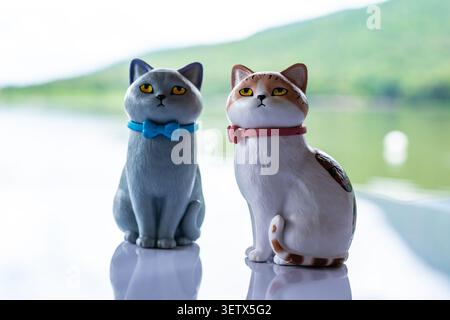 Chiang Mai, Thailand - 22 June 2026 : Gray and calico cat figurines with blue bow tie and pink collar sitting on reflective surface with unimpressed Stock Photo