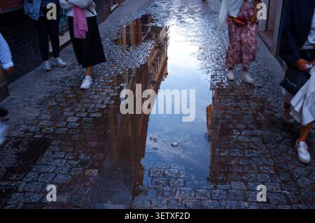 A rain puddle on the cobble streets of Seville Spain reflecting buildings as people walk by avoiding water. Stock Photo