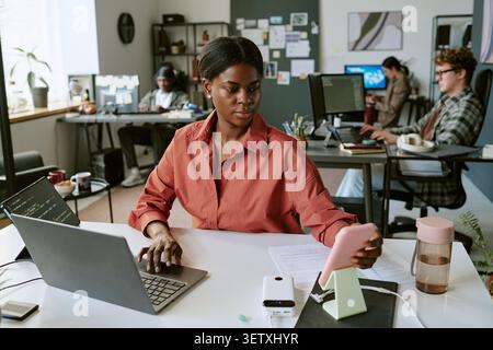 Black Woman Using Smartphone and Laptop in Modern Office Stock Photo