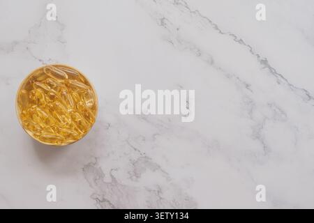 Top view of a small round wooden bowl filled with golden fish oil gel capsules placed on the left side of a white marble stone background with copy sp Stock Photo