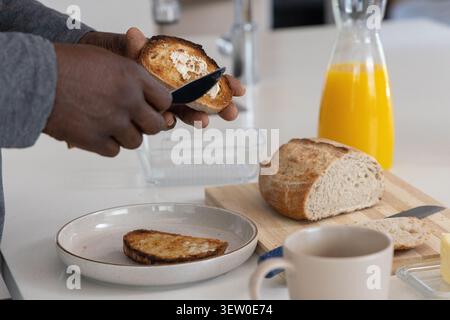 Mature adult male holding toast and spreading butter with knife on kitchen counter, copy space Stock Photo