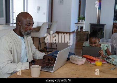 African American father in beige shirt working on laptop at dining table while child using tablet Stock Photo