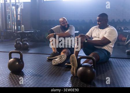 Two African men sitting on gym floor in sportswear chatting near kettlebell with trackers Stock Photo