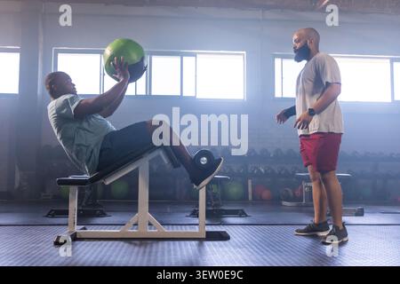 African American male trainer coaching client doing green med ball twist on incline bench at gym Stock Photo