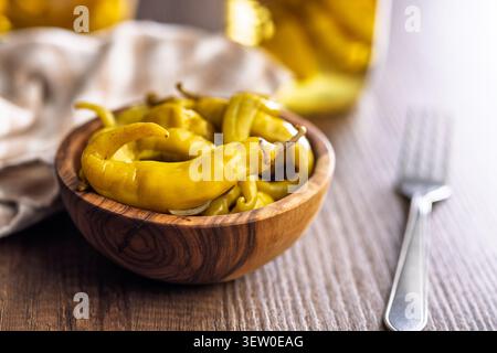 Bright pickled yellow banana peppers arranged in a wooden bowl on a rustic table, with a jar and fork in soft focus; flavorful, tangy appetizer or con Stock Photo