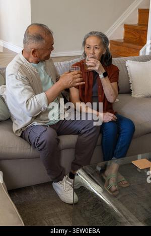Senior Asian couple sitting on sofa in living room, holding clear drinking glasses, smiling Stock Photo
