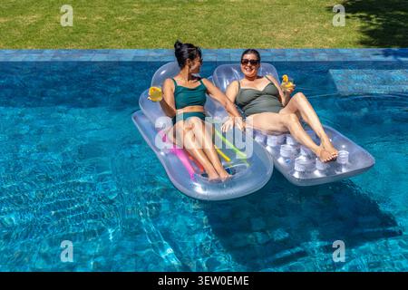 Two women reclining on loungers in backyard pool in swimsuits holding yellow cups and pool noodles Stock Photo