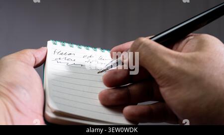 A POV shot of a male hand using a black pen to write on an open teal spiral bound notebook page, showing lined paper and details of fingers, set again Stock Photo