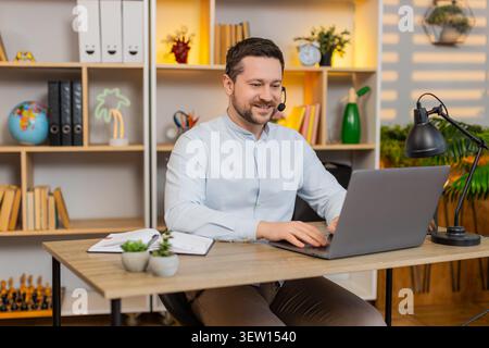 Young man on home table smiles in headset and listens to client on laptop while answering polite support call. Adult guy on home table works as remote operator giving help and service online each day. Stock Photo