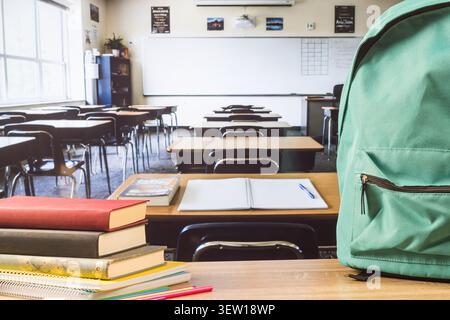Turquoise backpack, stacked books, notebooks and colored pencils on a wooden desk in an empty classroom, ready for back to school. Stock Photo