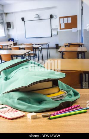 Open turquoise backpack packed with books and notebooks on a classroom desk, with calculator, eraser, sharpener and colored pencils nearby. Stock Photo
