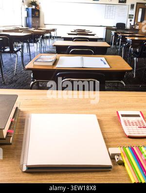 Empty classroom seen from a student desk with blank notebook, pink calculator, colored pencils, books, eraser and sharpener. Stock Photo
