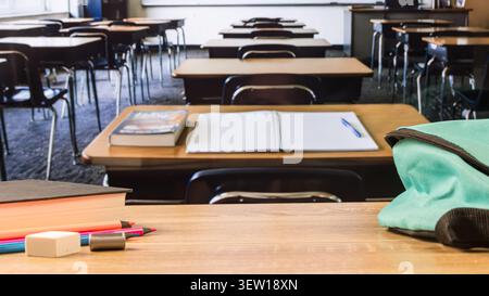 Wooden classroom desk with turquoise backpack, open notebook, book, colored pencils, eraser and sharpener, with empty desks in the background. Stock Photo