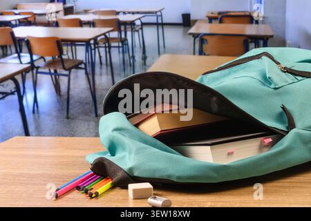 Open turquoise backpack filled with books and notebooks on a classroom desk, with colored pencils, eraser and sharpener in the foreground. Stock Photo