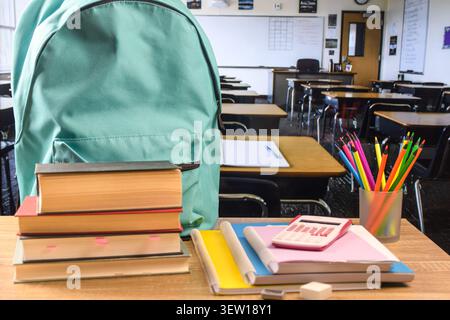 Turquoise backpack with books, notebooks, colored pencils, pink calculator, eraser and sharpener on a classroom desk. Stock Photo